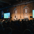 Vollbesetzter Konferenzsaal der UXDX EMEA 2026 mit Speaker auf der Bühne, Screens mit dem Konferenz-Logo und blau beleuchteter Industriehalle mit Backsteinwänden.